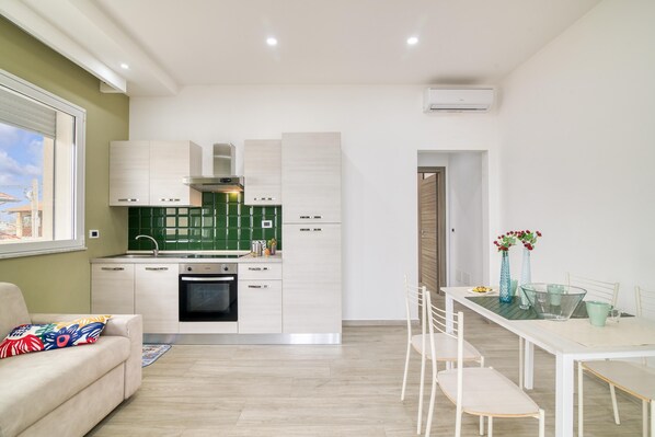 A modern kitchen with white cabinets, a built-in oven, and a green backsplash. There is a dining area with a white table and chairs, and a window with a view of buildings. - Corallo House 200 mt From the sea (Augusta)