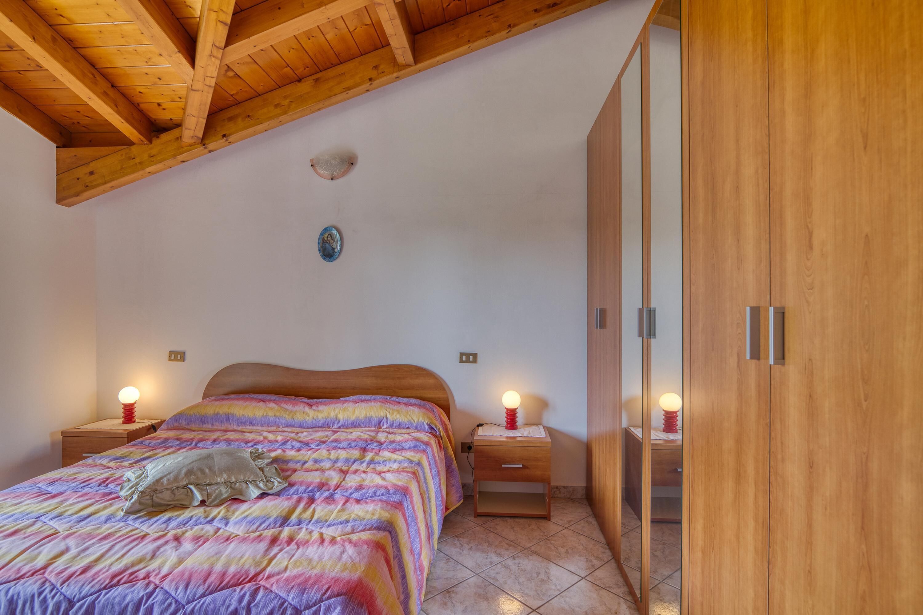 A bedroom with a wooden ceiling, a bed with a colorful blanket, two bedside tables with lamps, and a wooden wardrobe.