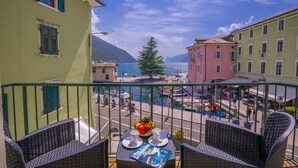 Un balcon agrémenté de meubles en osier, une table avec un panier de fruits et une vue sur une ville côtière, avec ses bâtiments et ses bateaux.