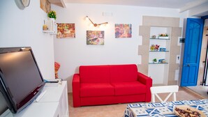A living room with a red sofa, a white chair, a TV on a white cabinet, and a table with a patterned tablecloth. - Casa Demetra 200m From sea (Syracuse)