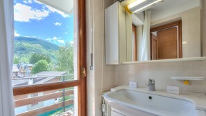 Une salle de bain dotée d’une grande fenêtre, d’un lavabo blanc, d’un miroir et donnant sur un balcon avec une balustrade en bois.