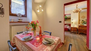 Un área de comedor con una mesa dispuesta para dos personas, una ventana con cortinas, una araña y vista a otra habitación.
