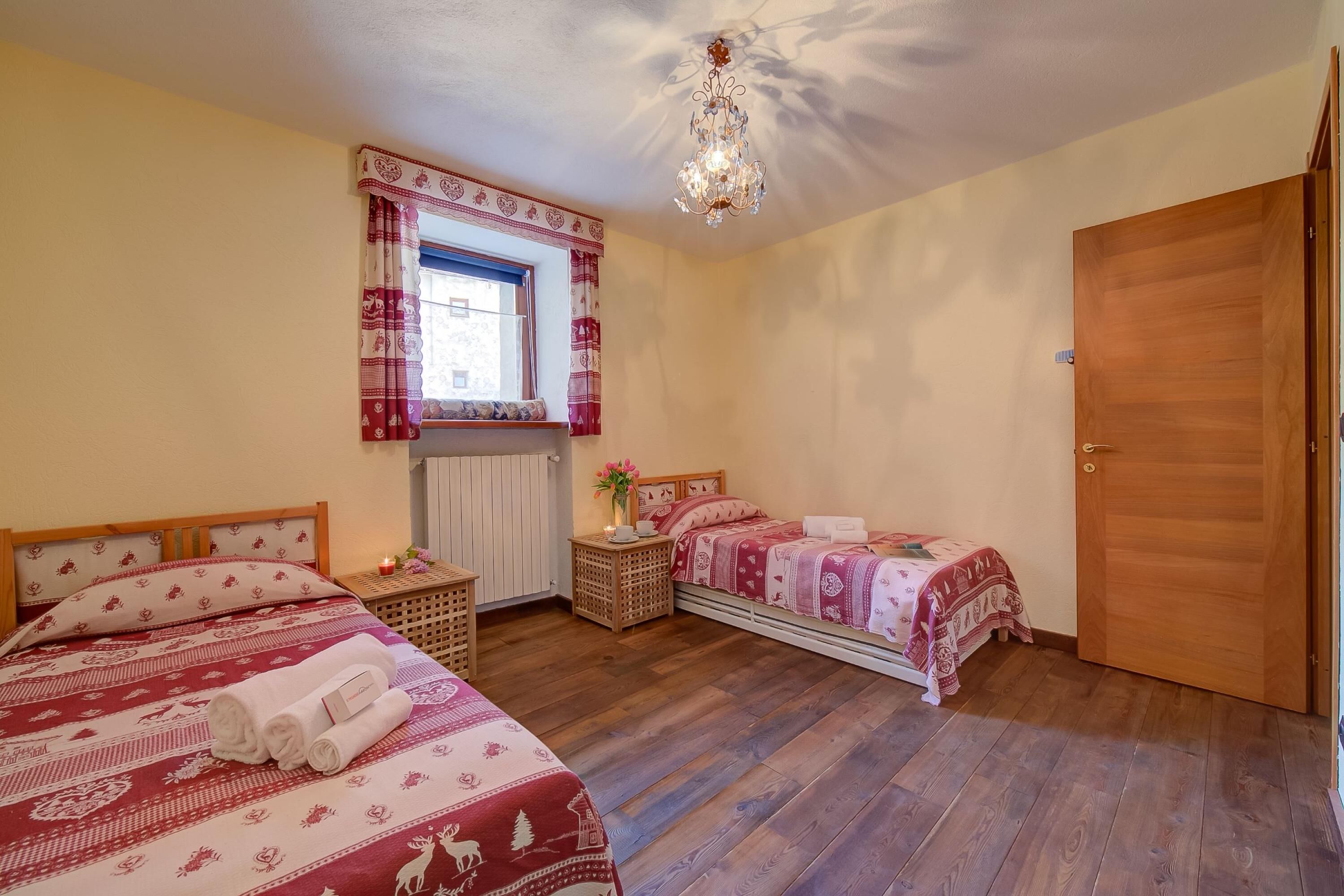 Un dormitorio con dos camas, una puerta de madera, un ventanal con cortinas, un candelabro y una mesita de noche con lámpara.