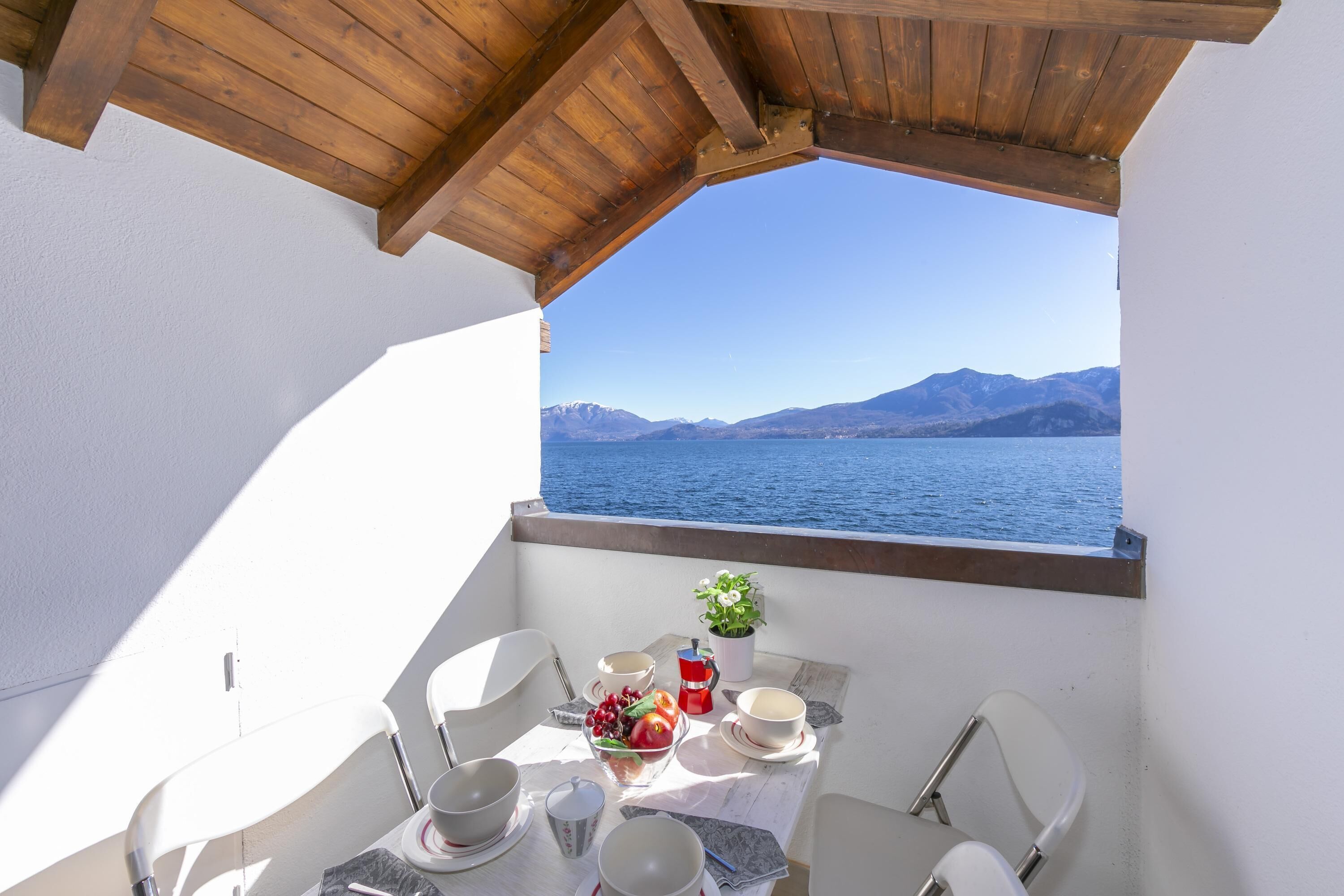A balcony with a wooden ceiling, white walls, and a table set for two with bowls, cups, and a fruit bowl. A potted plant is on the table, and a view of the sea and mountains is visible through the window.