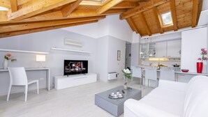 A modern living room with a TV, white furniture, and a kitchen in the background. - Blu Lake Apartment on Ghiffa Beach (Ghiffa)