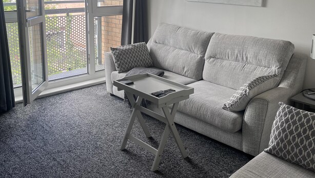 A living room with a grey sofa, a small table, and a large window with a view of a brick building. - 2 Bed Flat - Sleeps 4 - Pets - Parking - Wifi (Edinburgh)