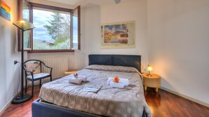 Ein Schlafzimmer mit einem großen Bett, einem Sessel, einer Stehlampe, einem Nachttisch mit Lampe sowie einem Fenster mit Blick auf Bäume und Gebäude.