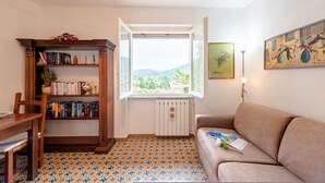 A cozy living room with a wooden bookshelf, a brown sofa, a patterned floor, and a window offering a view of greenery. - Il Cortiletto di Marina (Brovello-Carpugnino)