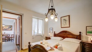 A bedroom with a wooden bed, a chandelier, a painting, and a dining area visible through an open door. - Il Cortiletto di Marina (Brovello-Carpugnino)