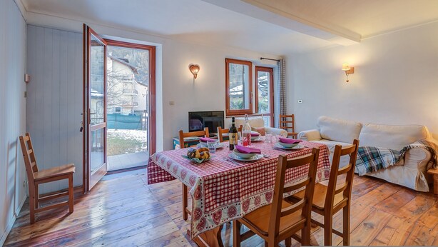 A dining area with a table set for four, wooden chairs, a sofa, and a TV in the background. - Appartamento Zia Anna Ski in Ski out (Sauze d'Oulx)