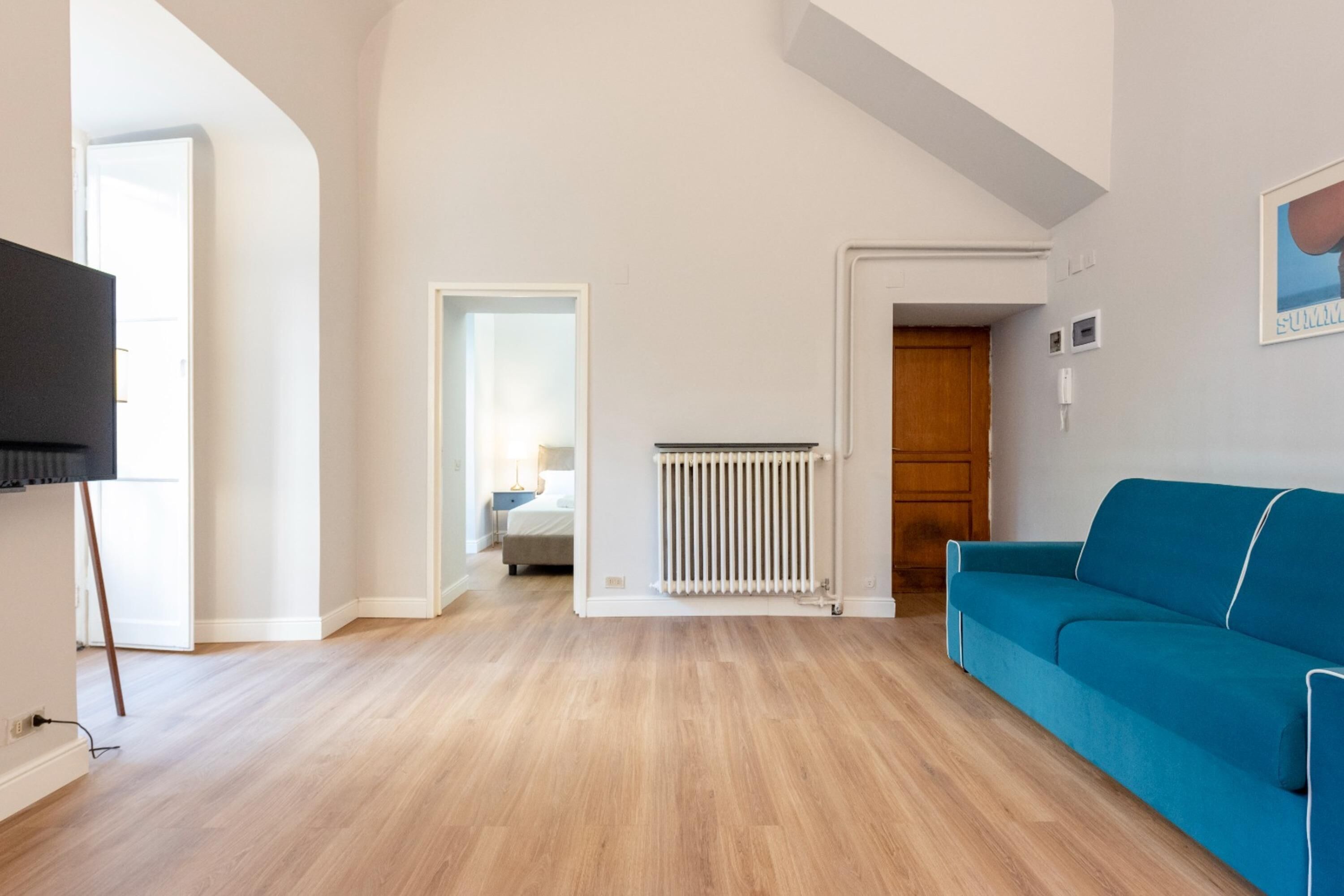 A spacious living room with a blue sofa, wooden flooring, a radiator, and a TV mounted on the wall.