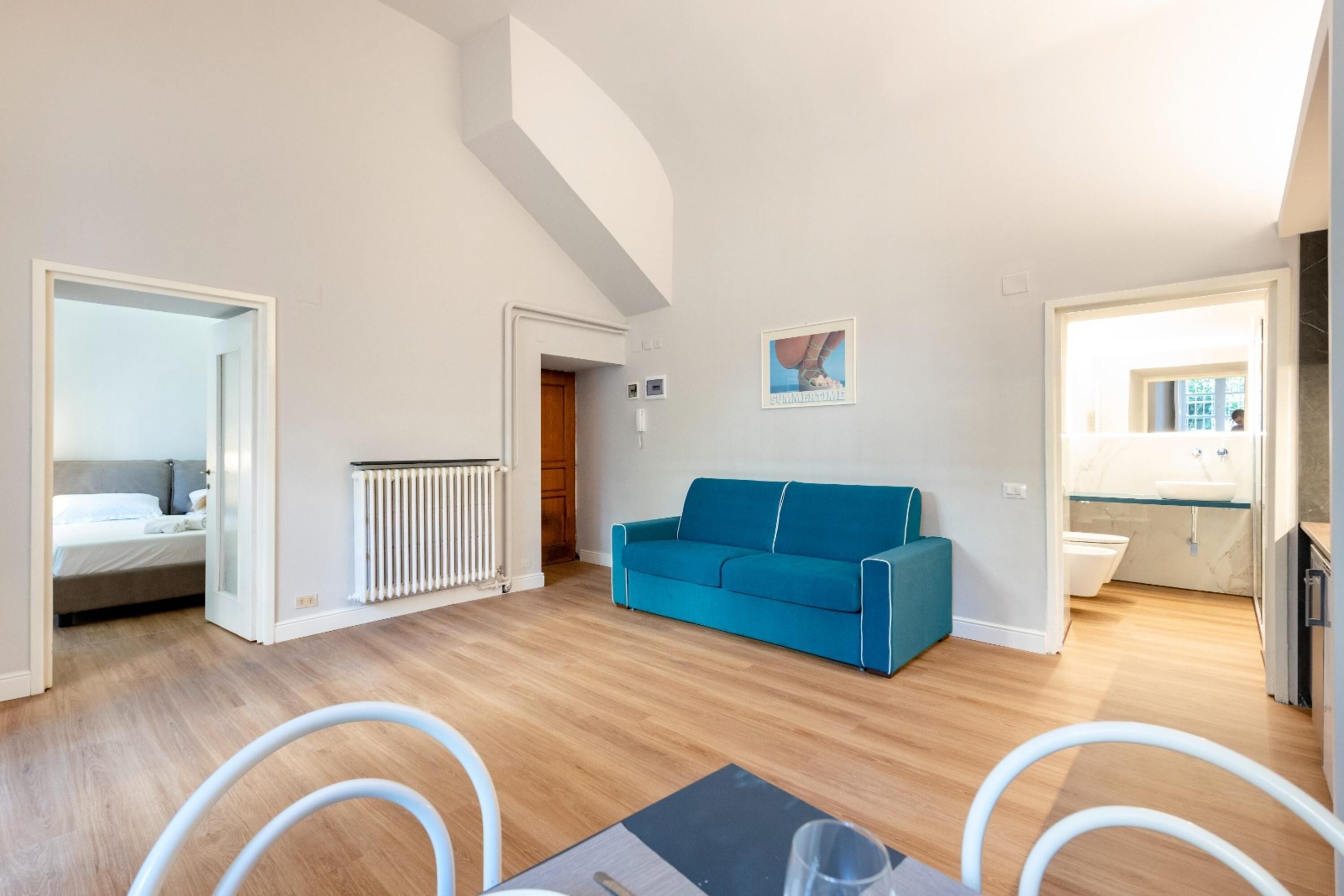 A modern living room with a blue sofa, a dining area with a table and chairs, and a kitchen visible through an open doorway.