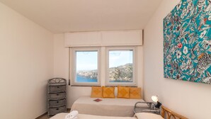Ein Schlafzimmer mit zwei Betten, Meerblick, einem Korbstuhl-Nachttisch und einem Gemälde an der Wand.