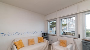 Ein Zimmer mit zwei beigefarbenen Sofas, jedes mit einem gelben Kissen, einem kleinen Tisch mit einer Lampe und Blick auf ein Gebäude durch ein Fenster.