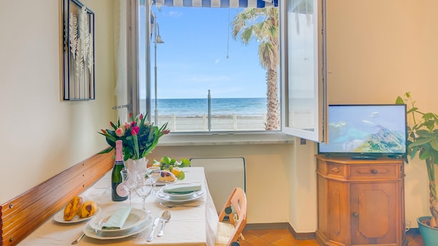 Un coin repas avec une table dressée pour deux personnes, une vue sur la plage et un téléviseur posé sur un meuble en bois.