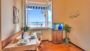 Un coin repas avec une table dressée pour deux personnes, une fenêtre donnant sur la plage et un téléviseur posé sur un meuble en bois.