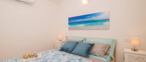 A bedroom with a bed, a nightstand, a lamp, a painting of a beach, and an air conditioner.