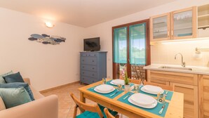 Zona pranzo con tavolo apparecchiato per due, un mobile azzurro, una televisione e una finestra con tende.