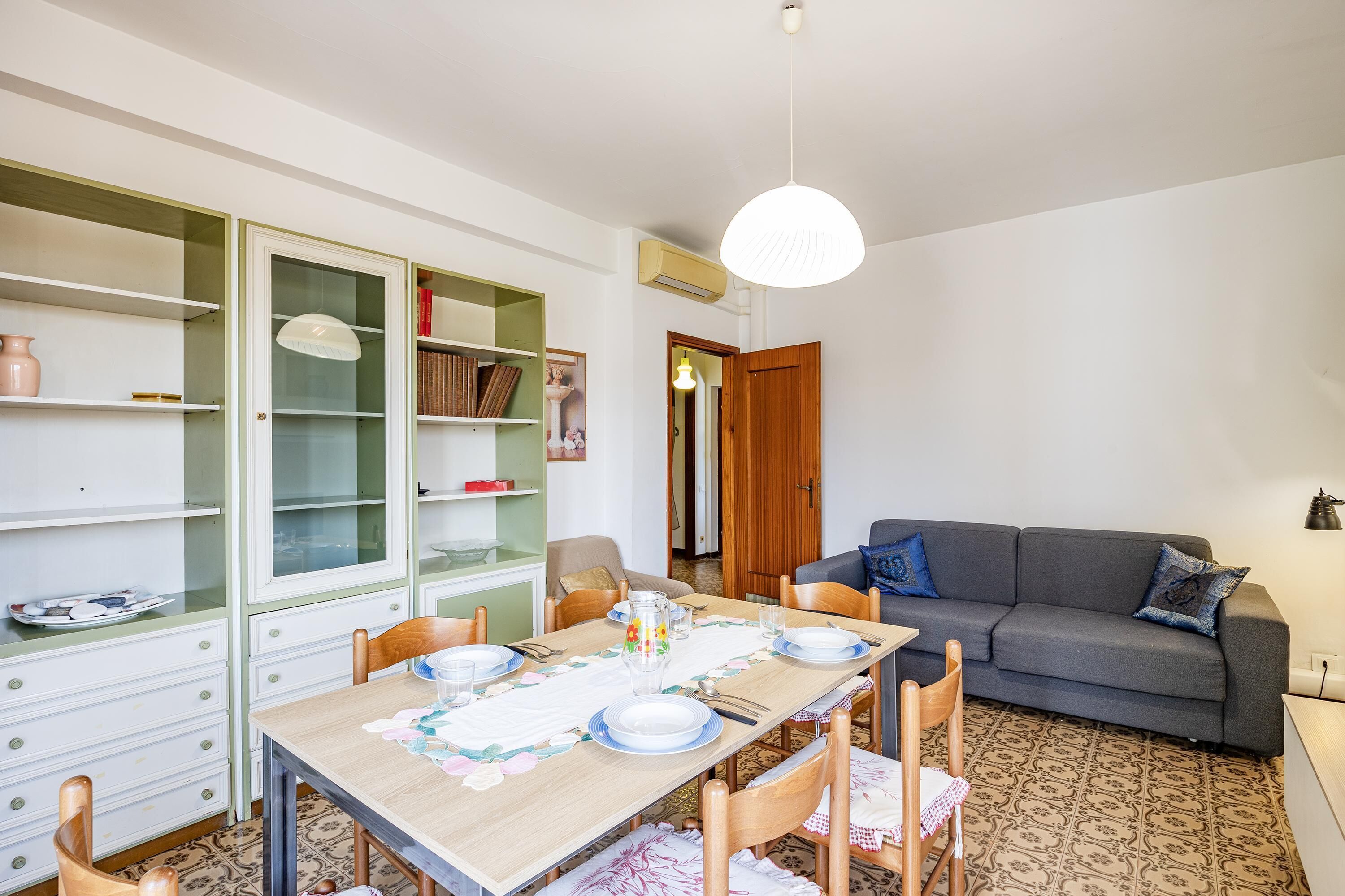 A dining area with a wooden table set for four, surrounded by chairs. There is a sofa and a cabinet with glass doors in the background.