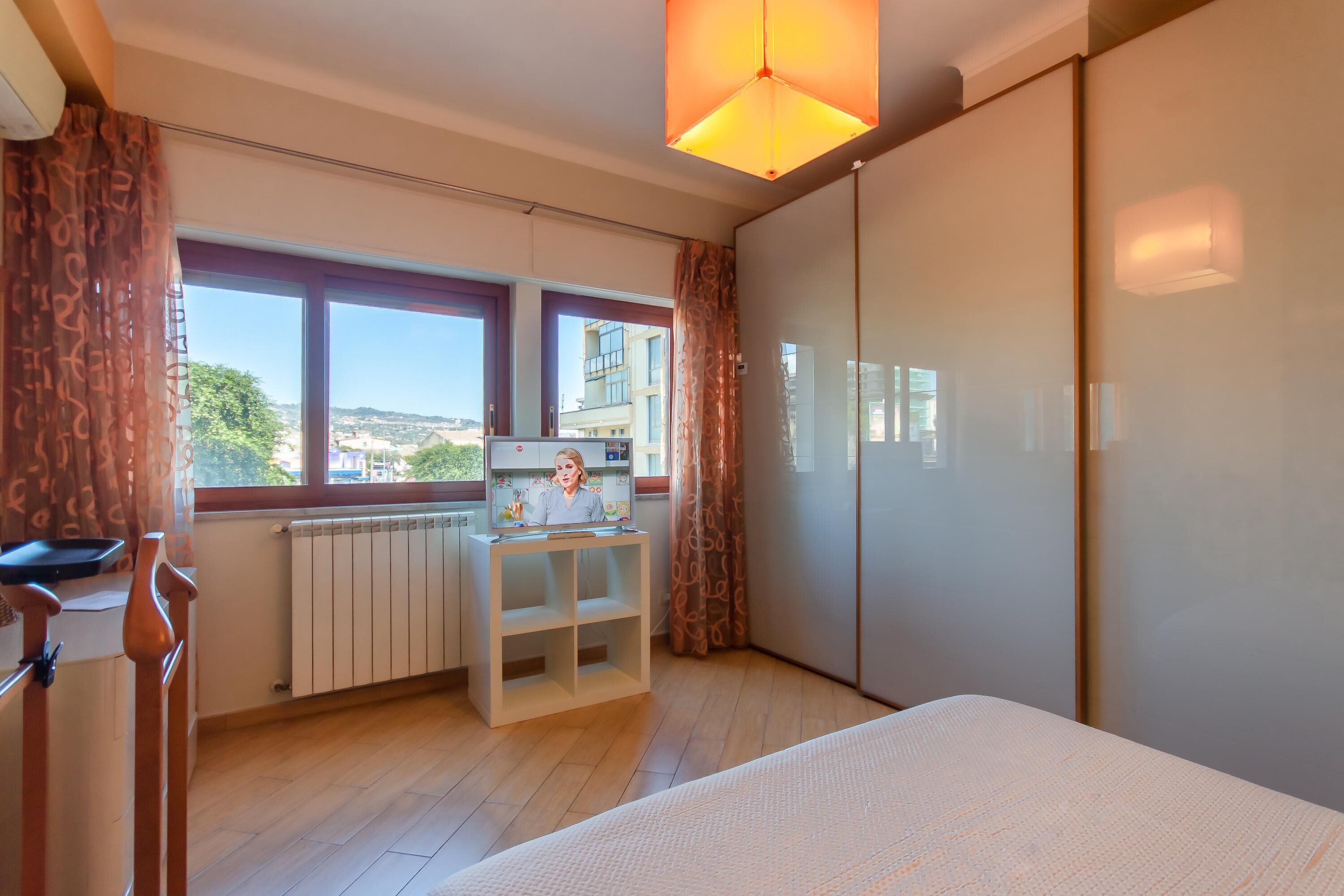 A bedroom with a bed, a desk with a chair, a radiator, a window with curtains, and a wardrobe.