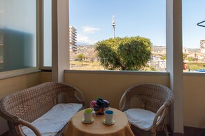 A balcony with wicker chairs, a small round table with a bowl of fruit and two cups, and a view of buildings and a tower in the distance. - Happy Apartment Nettuno Close To Sea (Catania)