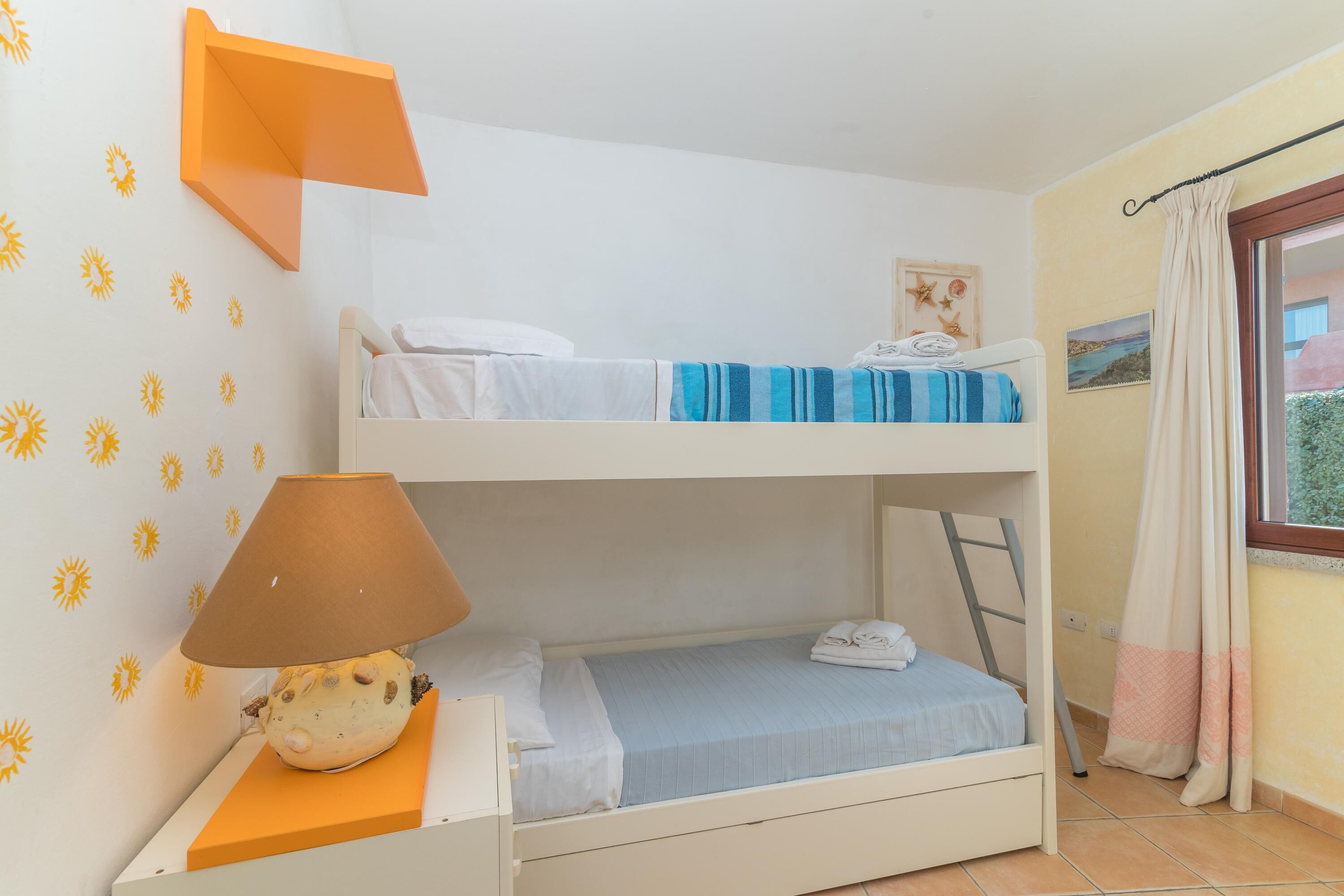 A bunk bed room with a lamp, a window with curtains, and a wall with sun decals.