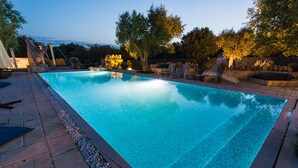 Et rektangulært swimmingpool med flisekant, omgivet af en stenbelagt terrasse og et par træer.