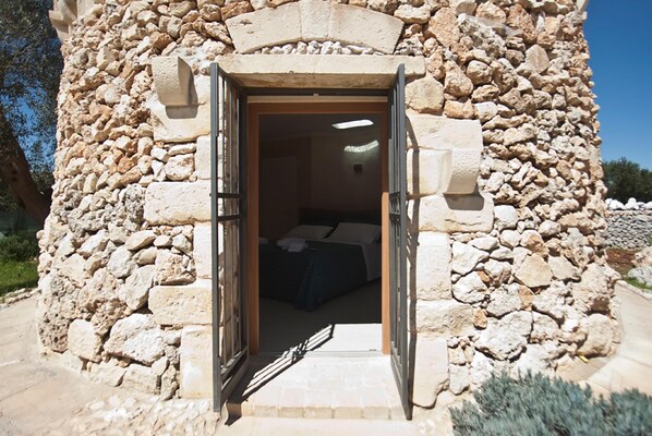 Un bâtiment en pierre avec une porte ouverte donnant sur une pièce où se trouvent un lit et une fenêtre.
