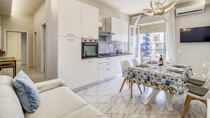 A modern kitchen with white cabinets, a dining area with a patterned tablecloth, and a living room with a sofa. - Bedo House Beach Front (Follonica)