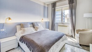 A bedroom with a bed, bedside lamps, a chair, a window with curtains, and a radiator. - Bedo House Beach Front (Follonica)