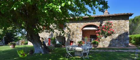 A stone building with a large tree in front, a swing, and a table with chairs.