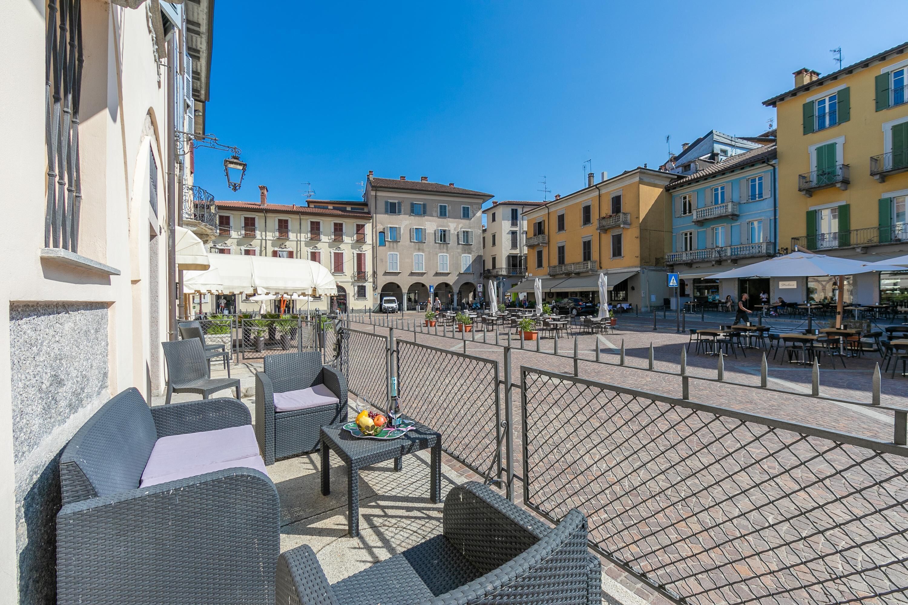 Una terrazza con mobili in vimini e un tavolo con frutta, che si affaccia su una piazza con edifici e tavoli all'aperto.