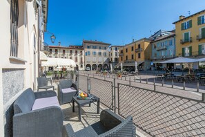 Une terrasse dotée de meubles en osier et d’une table garnie de fruits, donnant sur une place bordée de bâtiments et offrant des places assises en plein air.