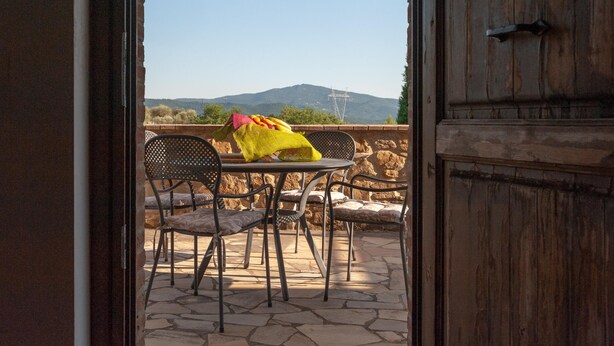Um pátio externo com mesa e cadeiras, piso de pedra e vista para uma montanha ao fundo.