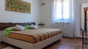 Quarto com cama, cabeceira, obra de arte na parede, janela com cortinas e porta de banheiro.