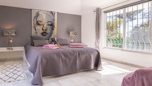 Une chambre moderne avec un grand lit, une vue sur la verdure par la fenêtre et une œuvre d’art abstraite au mur.