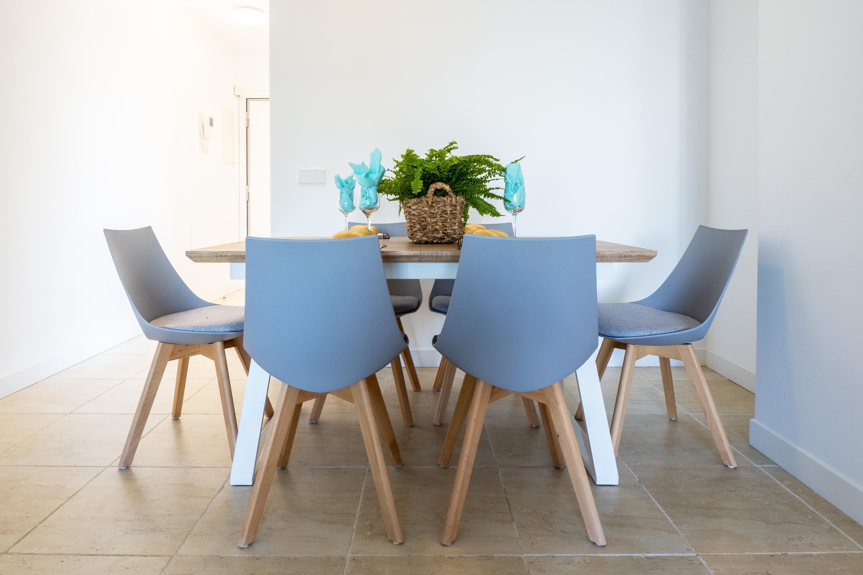 A dining area with a round table, four blue chairs, a woven basket with a plant, and a small wooden shelf.