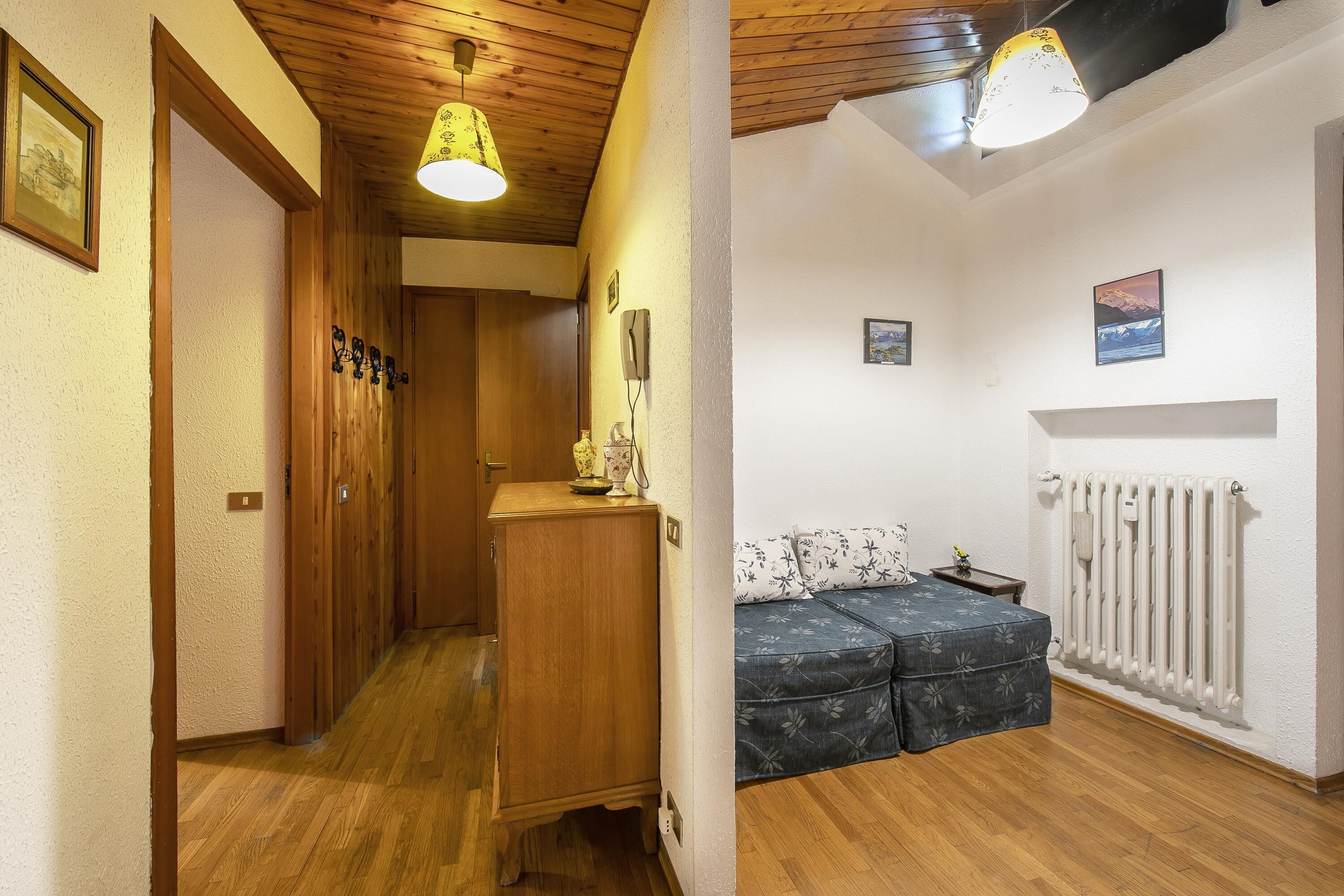 A hallway with wooden flooring and a wooden cabinet, leading to a room with a radiator, a bed, and a small table.