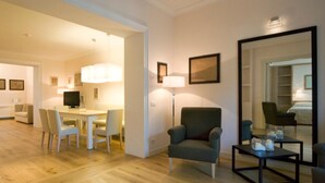 Moderni hotellihuone, jossa on puulattia, ruokailutila pöytineen ja tuoleineen, harmaa nojatuoli, musta sohvapöytä ja suuri peili, joka heijastaa huonetta.