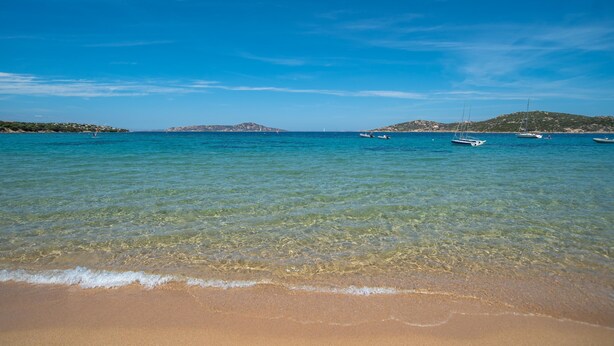 A beach with clear turquoise water, boats, and distant hills. - La Lampara House in Cannigione (Arzachena)