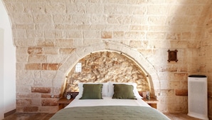 Une chambre avec un mur en pierre, un lit avec des coussins verts, une table de chevet en bois et un grand purificateur d’air blanc.