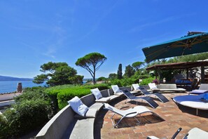 A sunlit outdoor seating area with lounge chairs and a covered table, overlooking a scenic coastal view. - Villa Ada in Ansedonia (Orbetello)