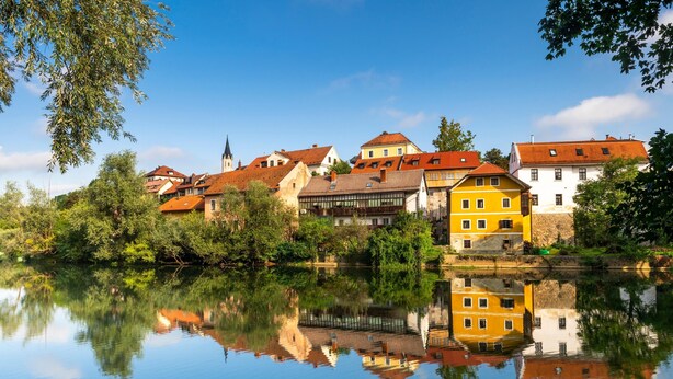 Une rivière paisible où se reflètent des bâtiments aux toits rouges et jaunes, le tout entouré de verdure.