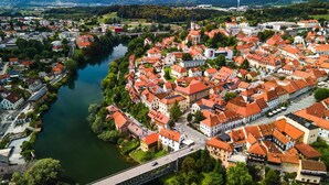Vue aérienne d’une ville traversée par une rivière, avec un pont, des bâtiments historiques aux toits rouges et un clocher d’église imposant.