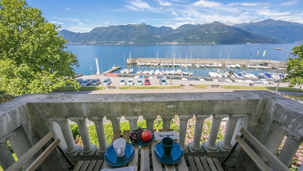 Blick von einem Balkon auf eine Marina mit vertäuten Booten, mit Bergen im Hintergrund und einem Tisch, gedeckt mit Tasse, Untertasse, Löffel und Obst.