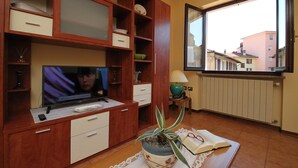 Un salon avec un téléviseur, des meubles en bois, une plante en pot et une fenêtre donnant sur des immeubles.