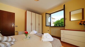 Une chambre à coucher avec un lit, une armoire en bois, une commode, une fenêtre donnant sur un paysage verdoyant et un vase rempli de fleurs.