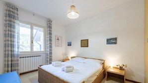 A bedroom with a large bed, a bedside table with a lamp, a wicker chair, and a view of the outdoors through a window. - Peaceful Apartment In Deiva Marina (Deiva Marina)