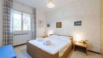 Una camera da letto con un letto grande, un comodino con una lampada, una poltrona in vimini e vista sull'esterno attraverso una finestra.