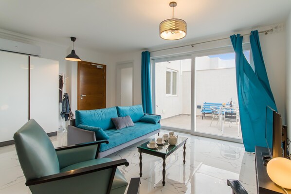 A modern living room with a blue sofa, a glass coffee table, and a view of the outside balcony. - Sunny Penthouse Modern 2 bdr - Sliema (Sliema)